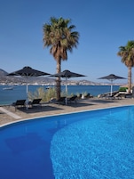 Una piscina con sillones de descanso y sombrillas, vista despejada del mar y una cadena montañosa a lo lejos.