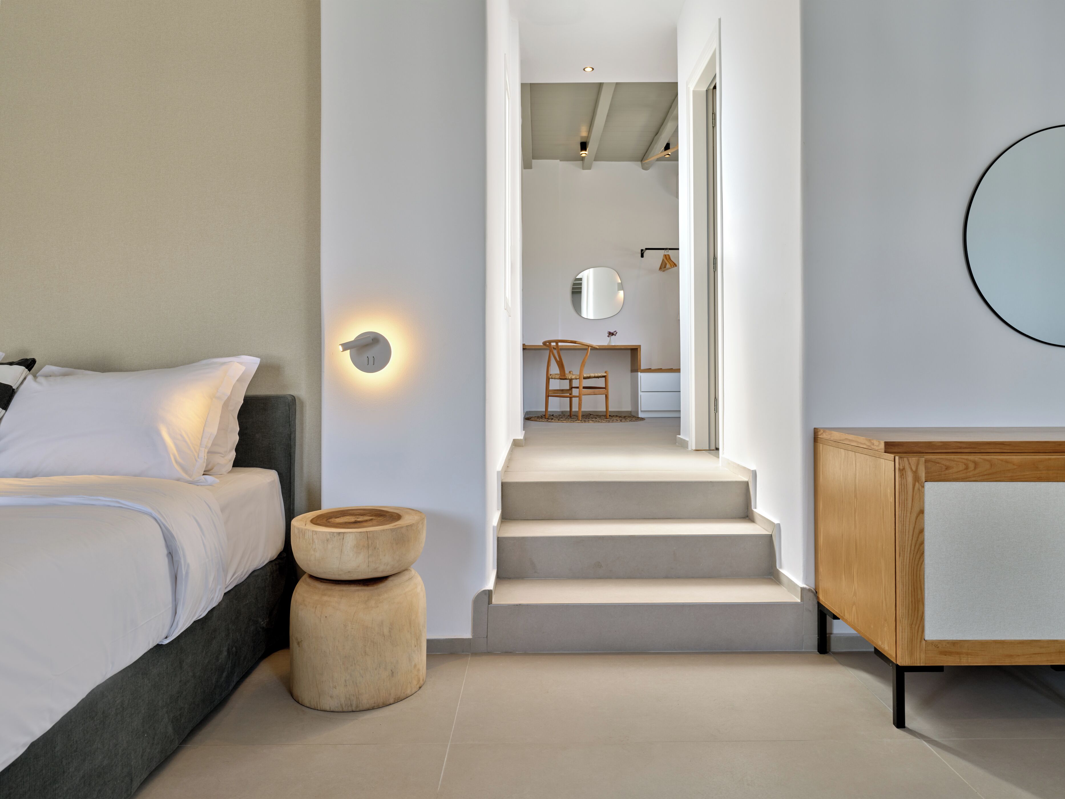 Une chambre à coucher moderne avec un lit, une table de chevet en bois, un miroir rond et un escalier menant à une salle de bain.