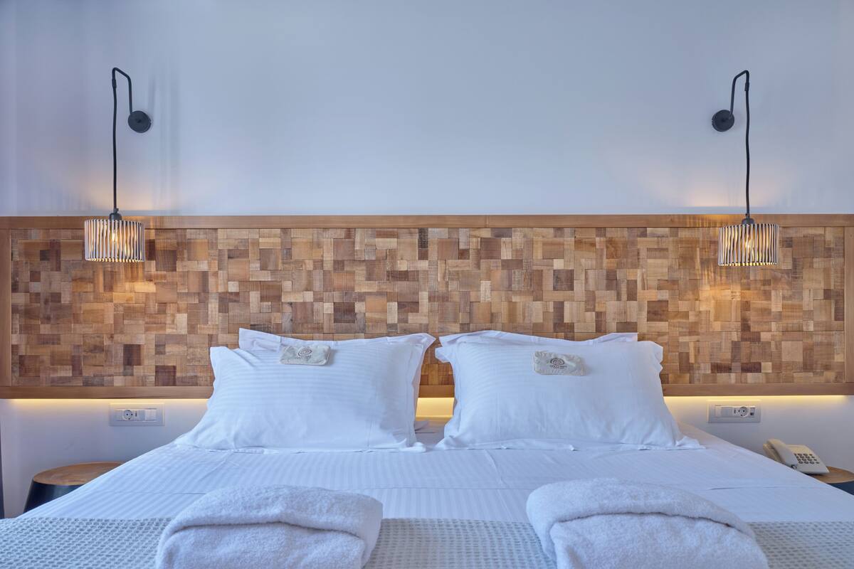 Habitación de hotel con cabecera de madera, dos almohadas blancas, una cama bien hecha, mesita de noche con lámpara y teléfono en la mesita.