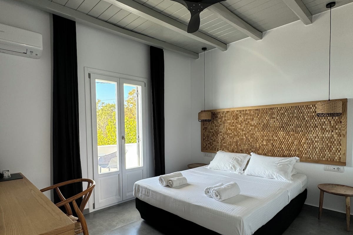 Una habitación de hotel moderna con una cama grande, un ventilador de techo, un cabecero de madera y vistas a través de un ventanal.