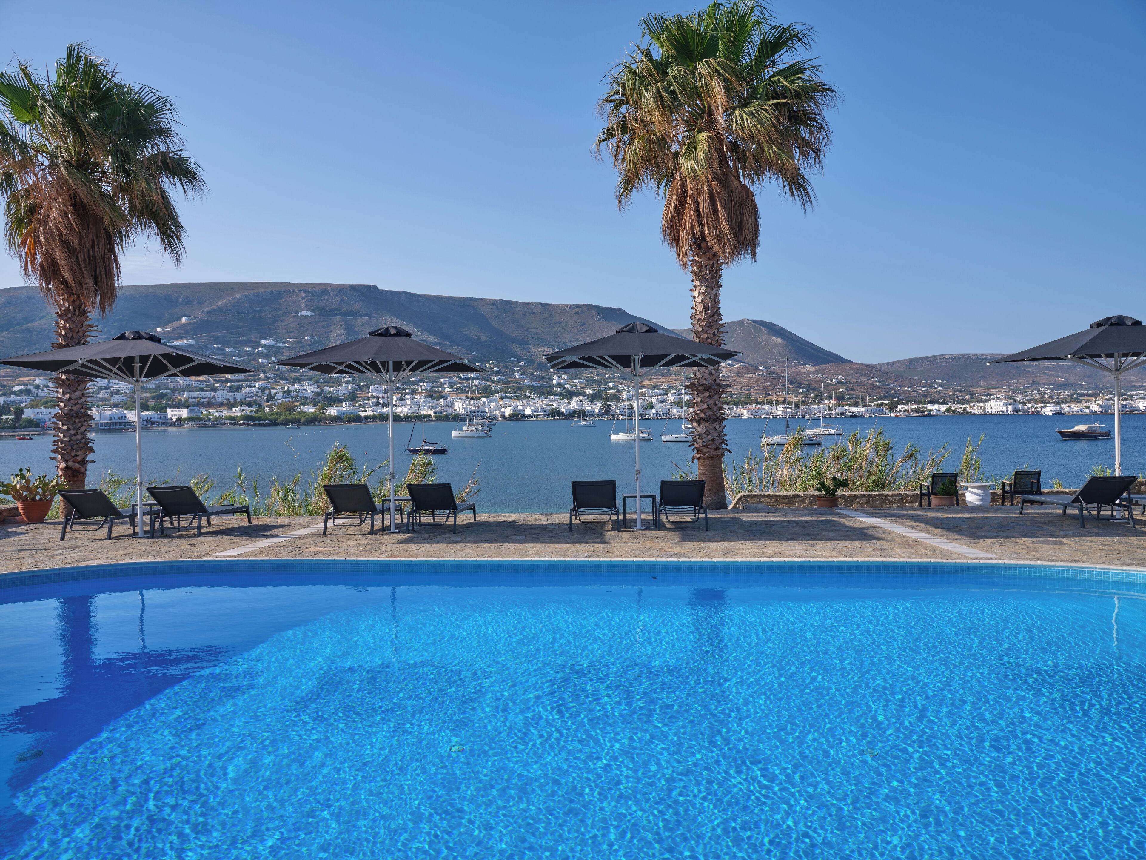 Zona junto a la piscina con sillones de descanso y sombrillas, con vistas a un cuerpo de agua donde se ven botes y un pueblo a lo lejos.