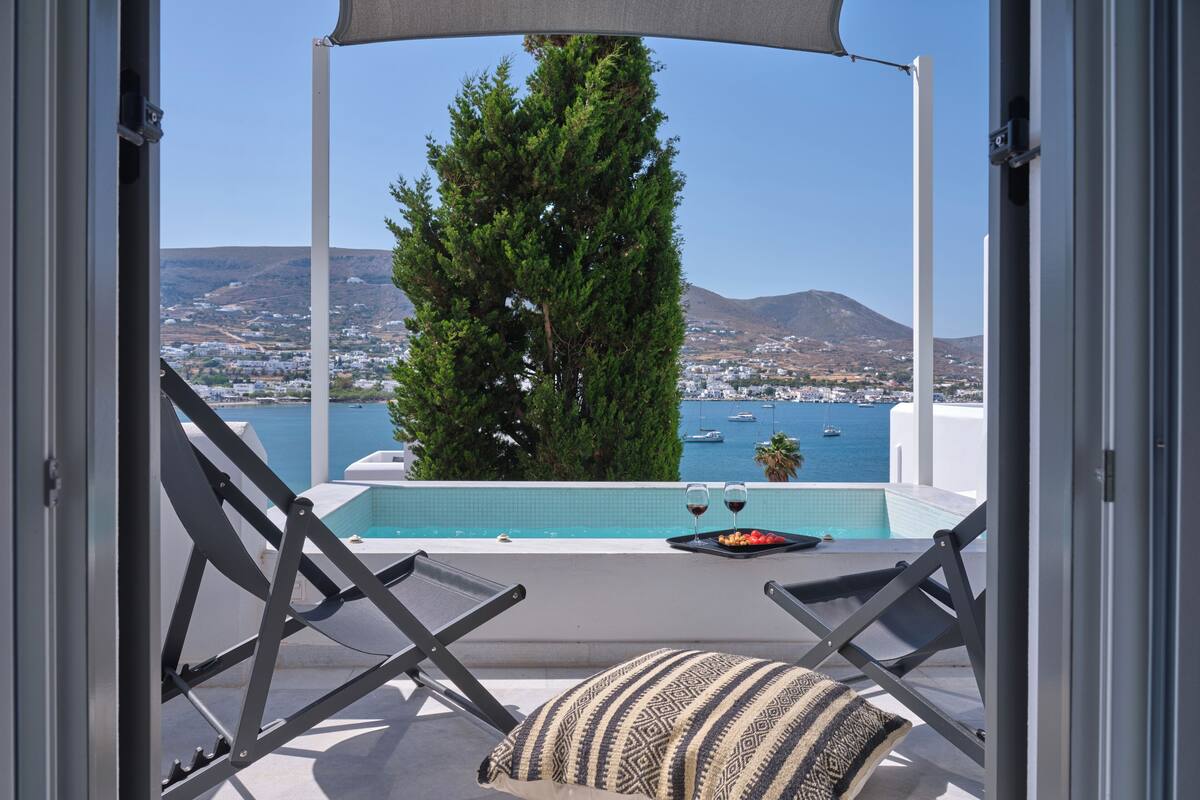 Zona de piscina con dos sillones de descanso y un cojín, con vistas a una masa de agua con barcos y un paisaje montañoso.