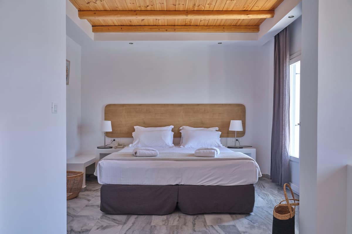 Una habitación de hotel moderna con techo de madera, una cama grande con ropa blanca, dos lámparas de noche y piso con diseño de mármol.
