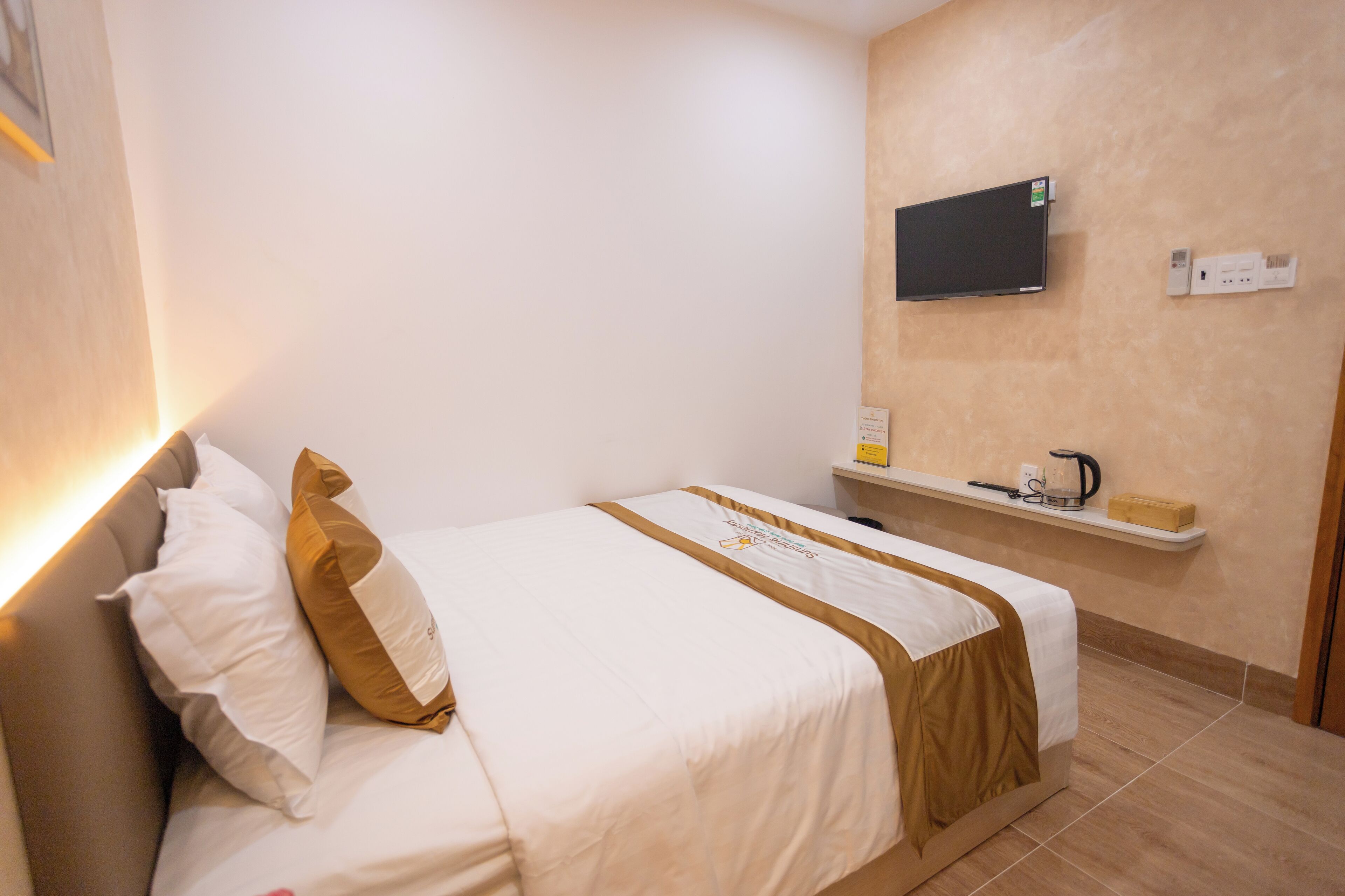 Ein ordentlich bezogenes Bett mit weißer und brauner Bettwäsche, ein flachbildfernseher an der Wand und ein kleines Regal mit Wasserkocher und einer Kiste.