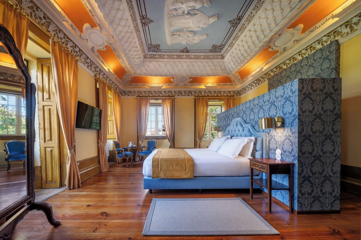 A luxurious bedroom with a large bed, a sitting area with chairs, a television, and ornate ceiling details.