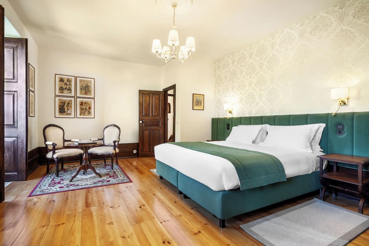 A bedroom with a large bed, a wooden floor, a dining table with chairs, and a chandelier.
