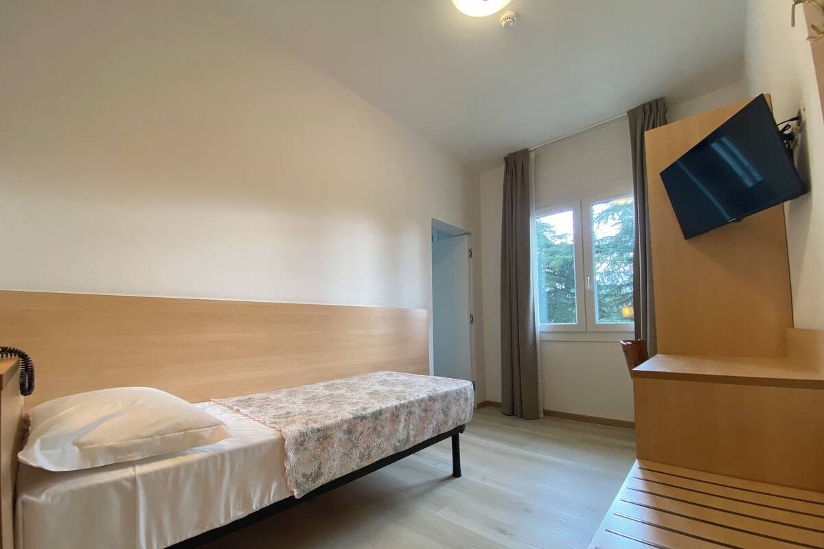 Uma cama de solteiro com cabeceira de madeira, mesa de cabeceira com luminária e televisão fixada na parede.