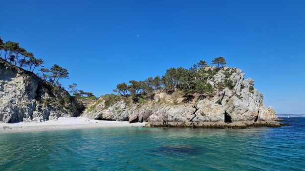 Eine felsige Küstenlandschaft mit klarem türkisfarbenem Wasser, einem Sandstrand und dichten Kiefern auf einer Klippe.