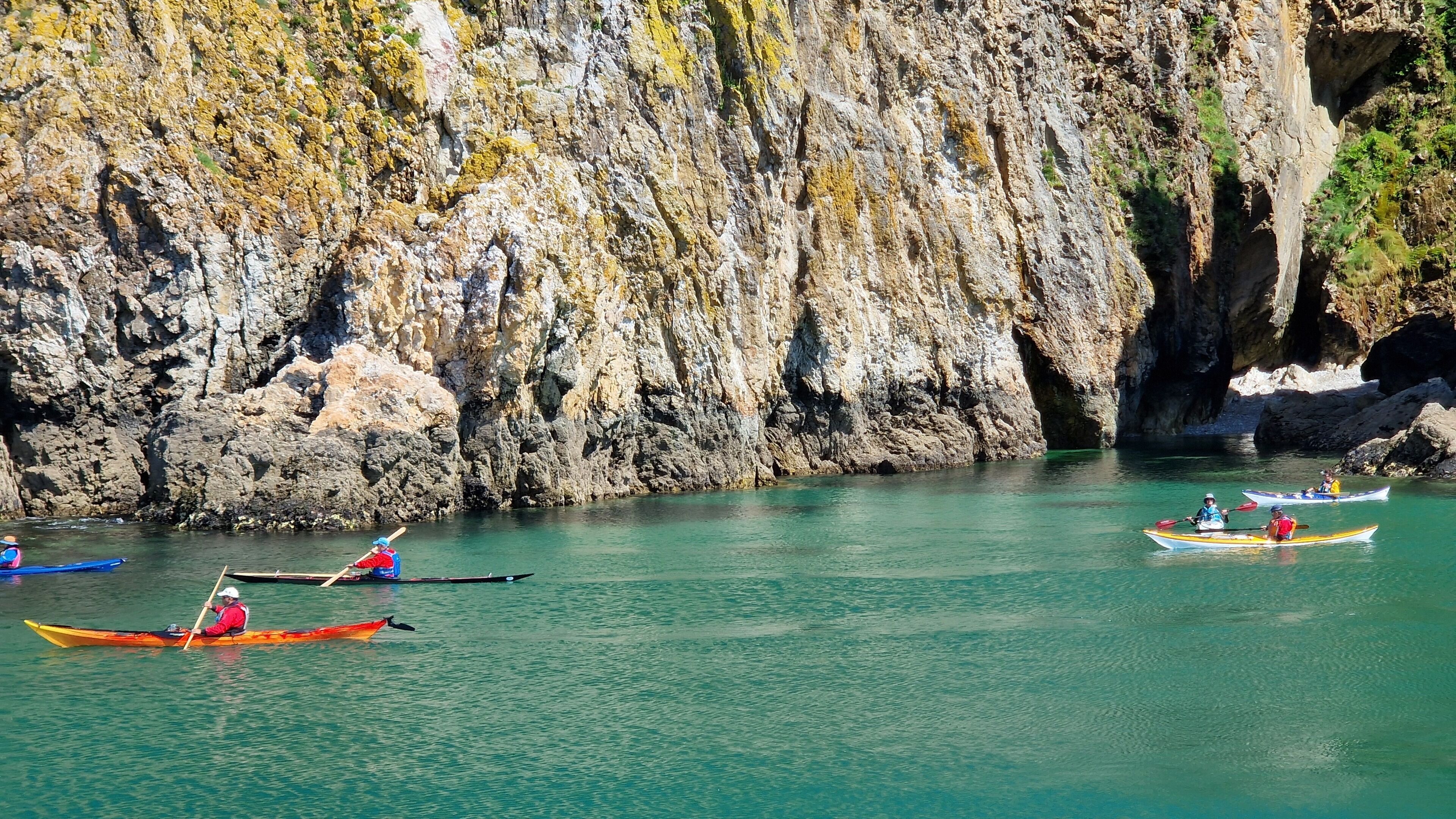 A group of kayaks on calm water with rocky cliffs in the background.