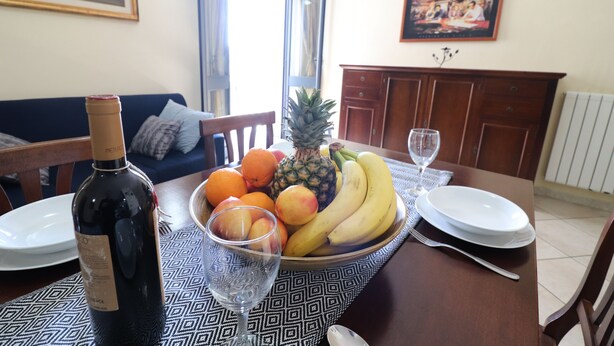 Een eettafel met een fles wijn, een glas en een schaal fruit met bananen, sinaasappels en een ananas.