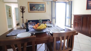 Een eettafel is gedekt met borden, bestek, een schaal fruit en een fles. Aan de muur hangt een schilderij, er staat een kapstok en er is een raam met gordijnen.