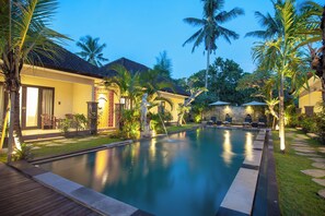 A swimming pool with a wooden deck, surrounded by palm trees and a building with a tiled roof. - d'Kamala Ubud by Prasi (Ubud)