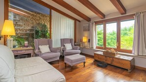 A cozy room with wooden beams, a stone wall, and large windows offering a view of greenery. There are two armchairs, a sofa, and a wooden side table with a lamp. - Son ArboÇ Gran - Villa With Private Pool (Algaida, Illes Balears)