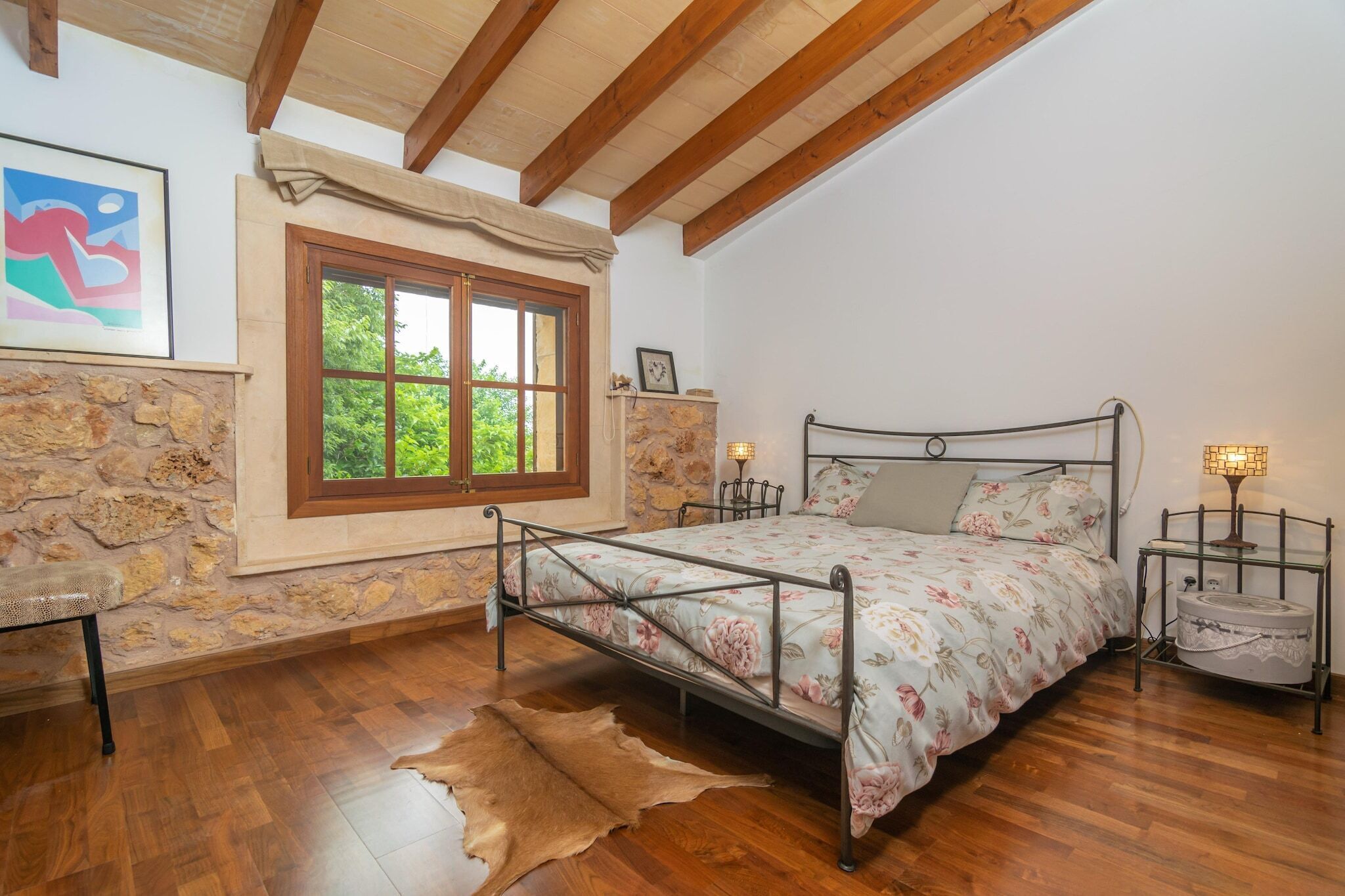 Quarto com parede de pedra, piso de madeira, cama com roupa de cama floral, um banco, janela com vista e mesa de cabeceira com abajur.