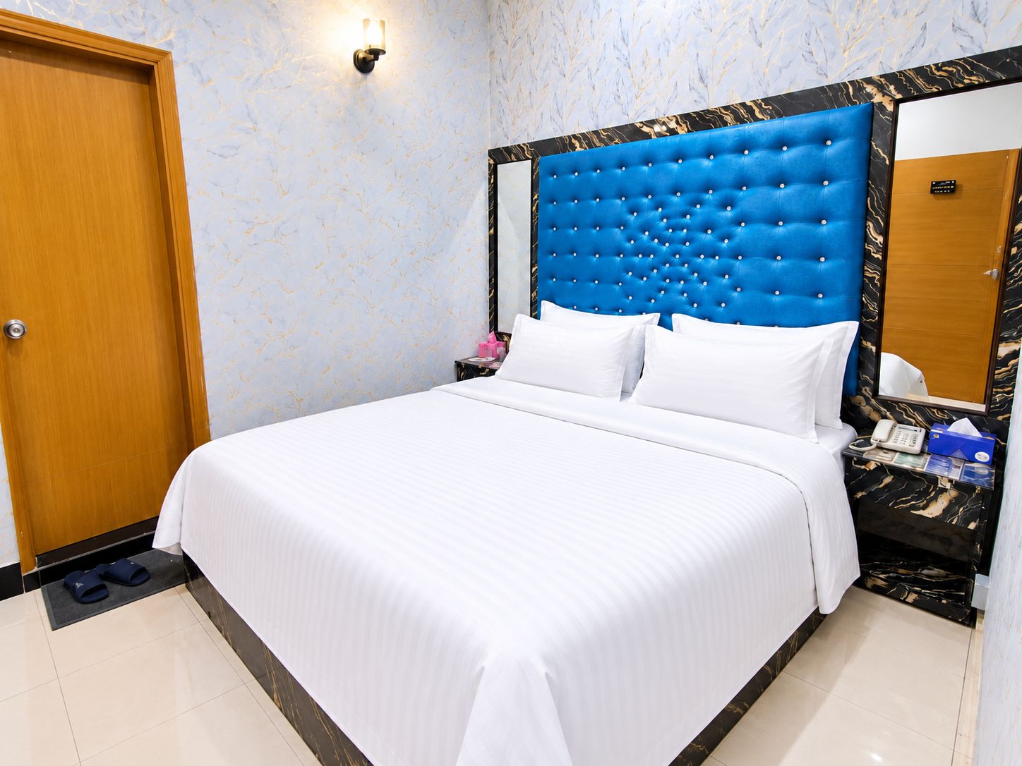 Ein Hotelzimmer mit einem großen Bett, einer Holztür, einer Wand mit Marmoroptik und einem blauen Kopfteil.