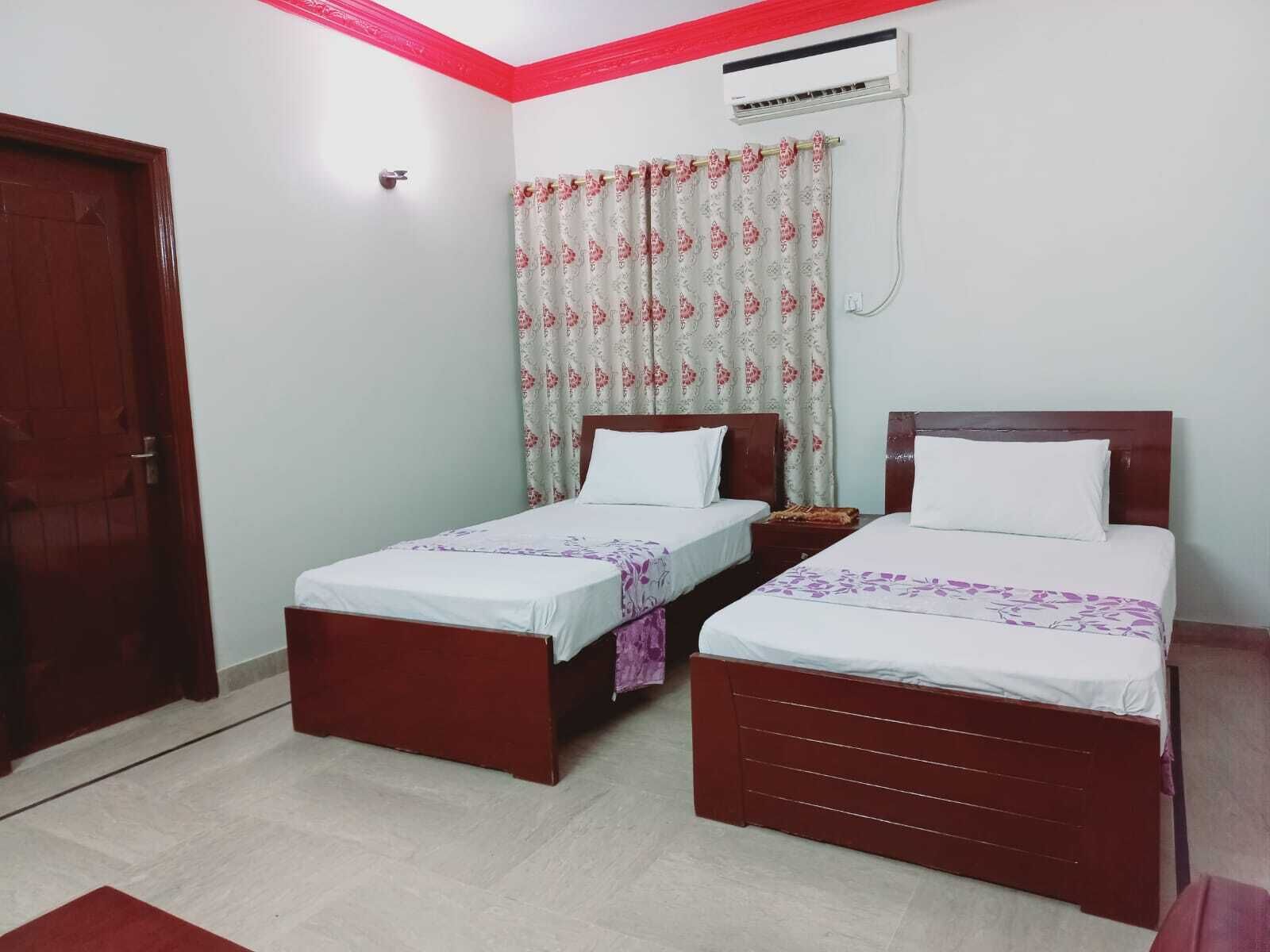 A room with two single beds, a wooden door, a red accent wall, and a patterned curtain.