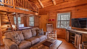 Un acogedor interior de cabaña con escalera de madera, un sofá, un sillón mecedor, un televisor y una chimenea.