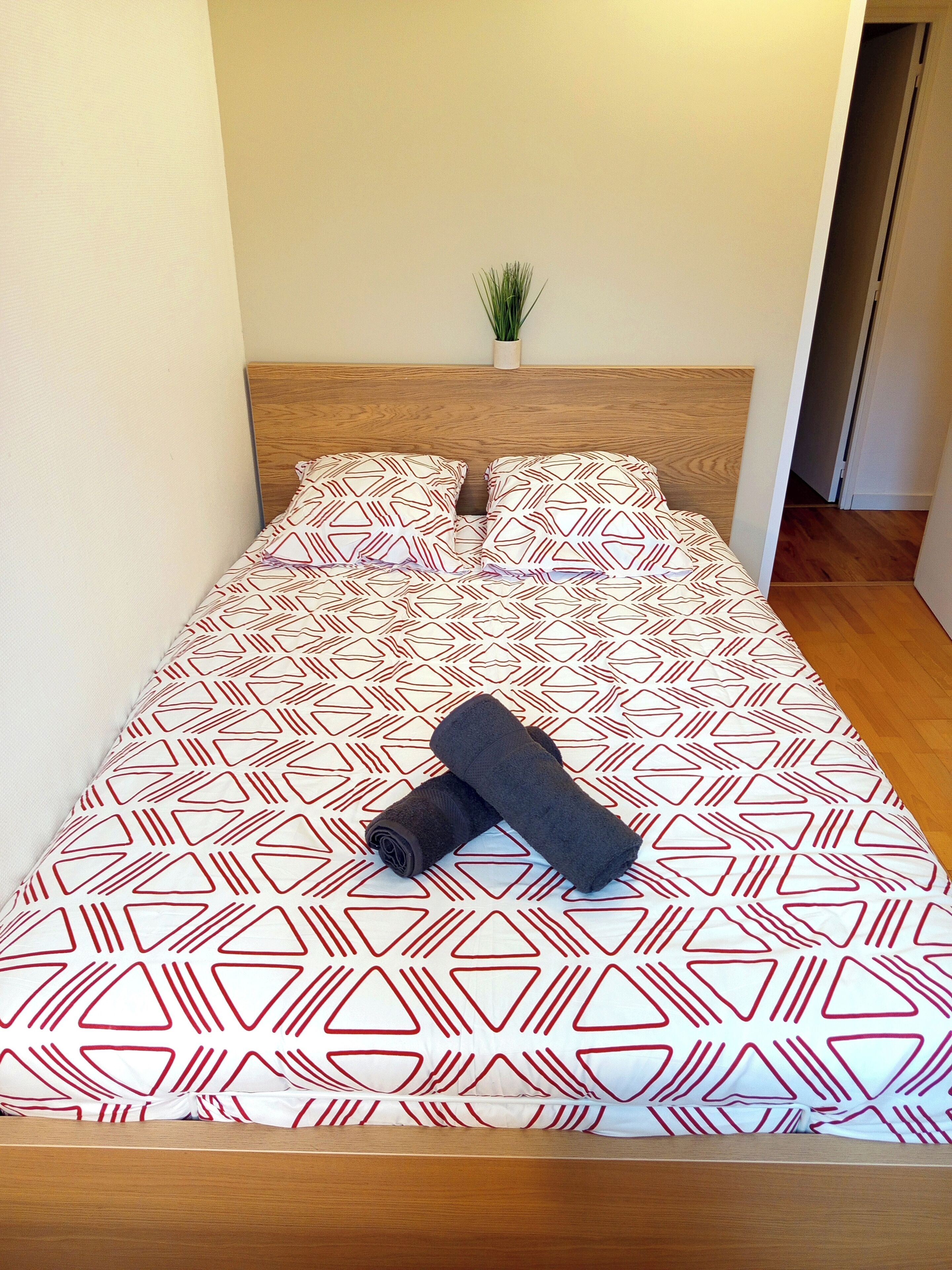 A neatly made bed with patterned bedding, a small potted plant on the headboard, and wooden flooring.