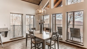 A dining area with a large table and chairs, large windows with views of trees and hills, and a hanging light fixture. - The Foxfire Cabin - Tree-top Getaway (Ponca)