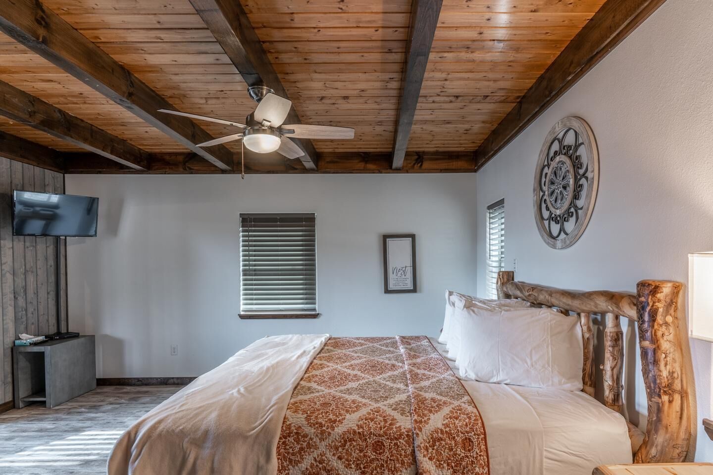 A bedroom with a wooden ceiling, a bed with a patterned bedspread, a ceiling fan, a television, and a window with blinds.