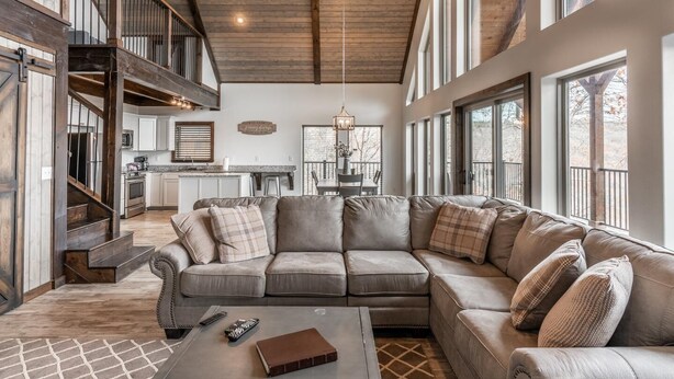 A spacious living area with a large L-shaped sectional sofa, a wooden staircase, and a kitchen in the background. - The Foxfire Cabin - Tree-top Getaway (Ponca)