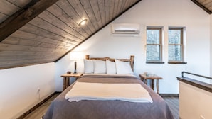 A bedroom with a sloped ceiling, a bed with a wooden headboard, two bedside tables, a nightstand, a lamp, and two windows. - The Wildwood Cabin - Tree-top Getaway (Ponca)