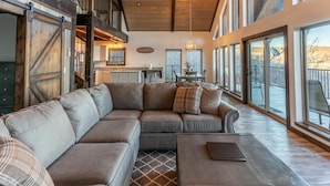 A spacious living room with a large sectional sofa, a coffee table, and a view of the outdoors through large windows. - The Wildwood Cabin - Tree-top Getaway (Ponca)