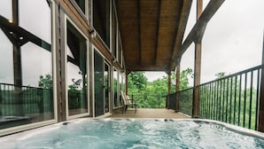 A hot tub on a wooden deck with glass railings, overlooking a lush green landscape. - The Wildwood Cabin - Tree-top Getaway (Ponca)