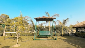 Un columpio verde en un área con césped, palmeras y una estructura de techo de paja al fondo.