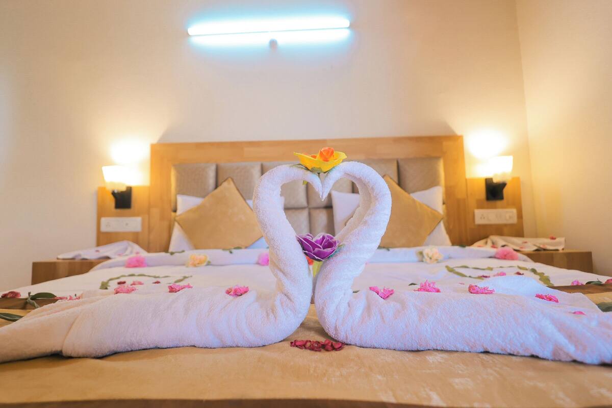 Habitación de hotel con una cama, adornada con dos toallas dobladas en forma de cisnes y flores, un cabecero de madera y dos lámparas de noche.