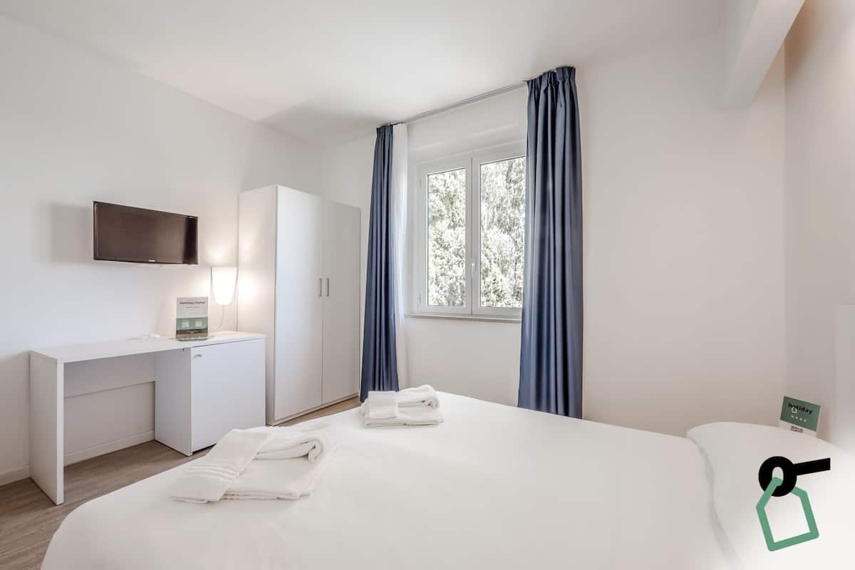A hotel room with a bed, a desk, a flat-screen TV, a telephone, and a window with curtains.