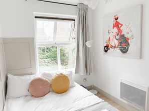 Un dormitorio con cama, ventana, radiador y un cuadro de una mujer en un patinete.