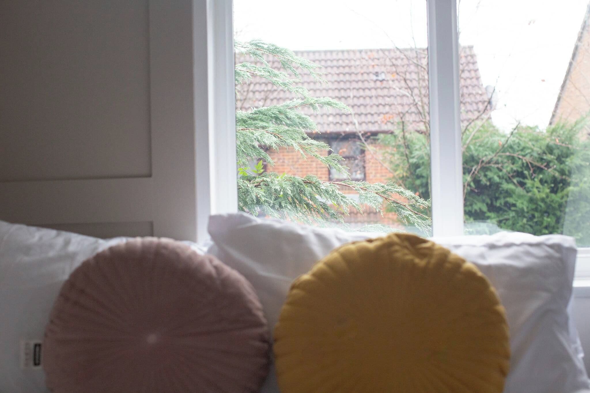 A bed with white pillows and a window with a view of a brick building and greenery.