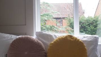 A bed with white pillows and a window with a view of a brick building and greenery.