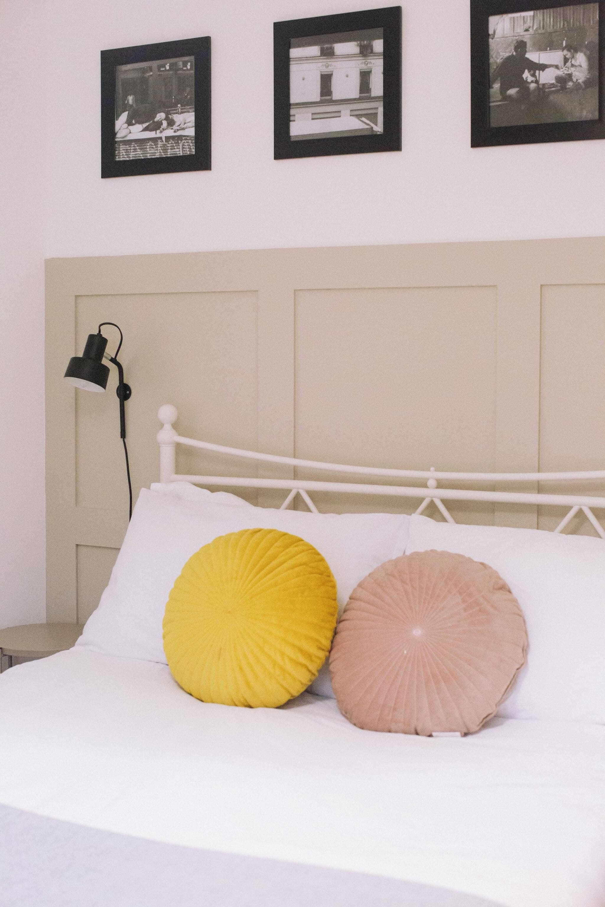 A bed with white bedding and two decorative pillows, a white headboard, and a wall-mounted lamp.