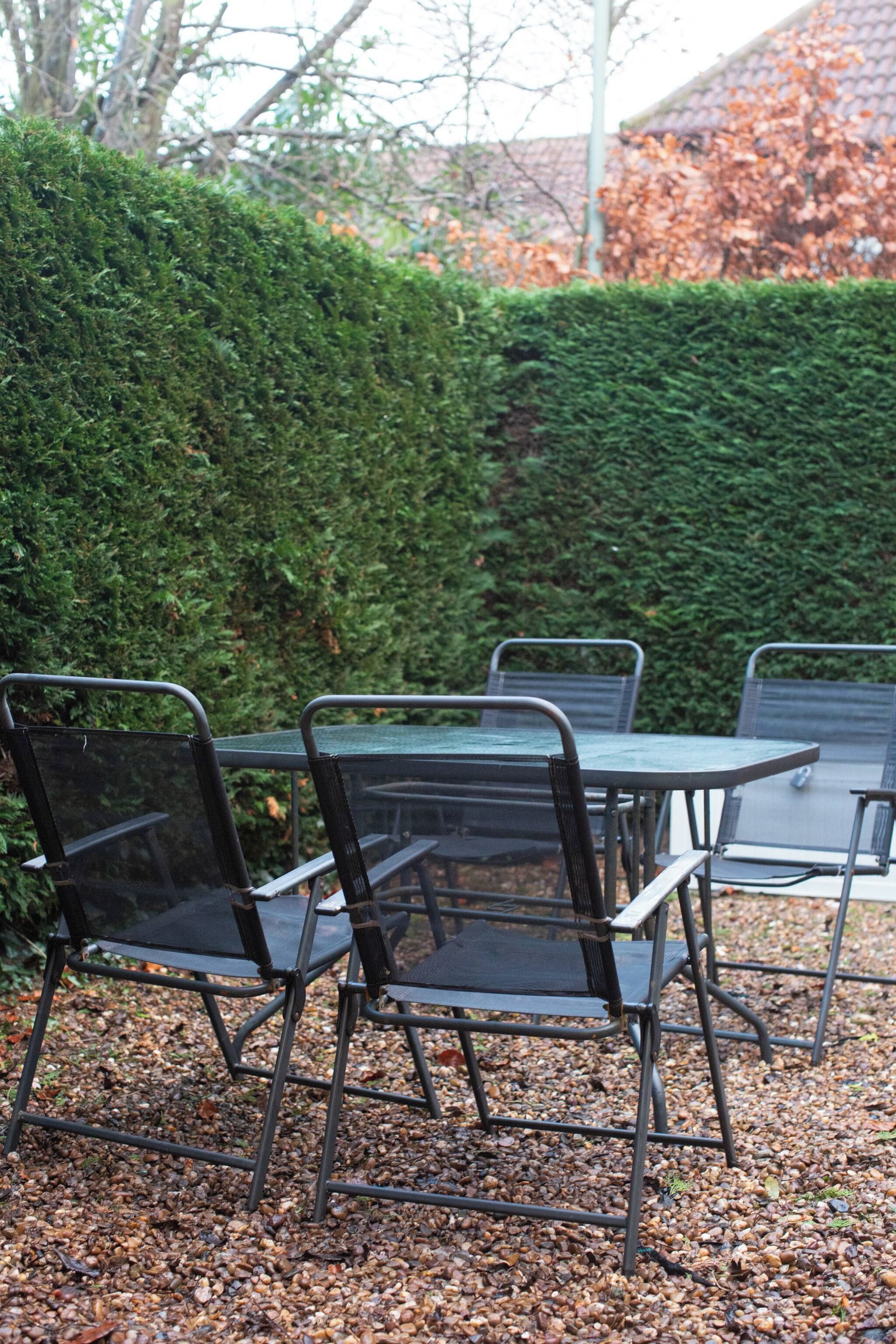 Een terras met metalen tafels en stoelen, omgeven door een haag en een grindpad.