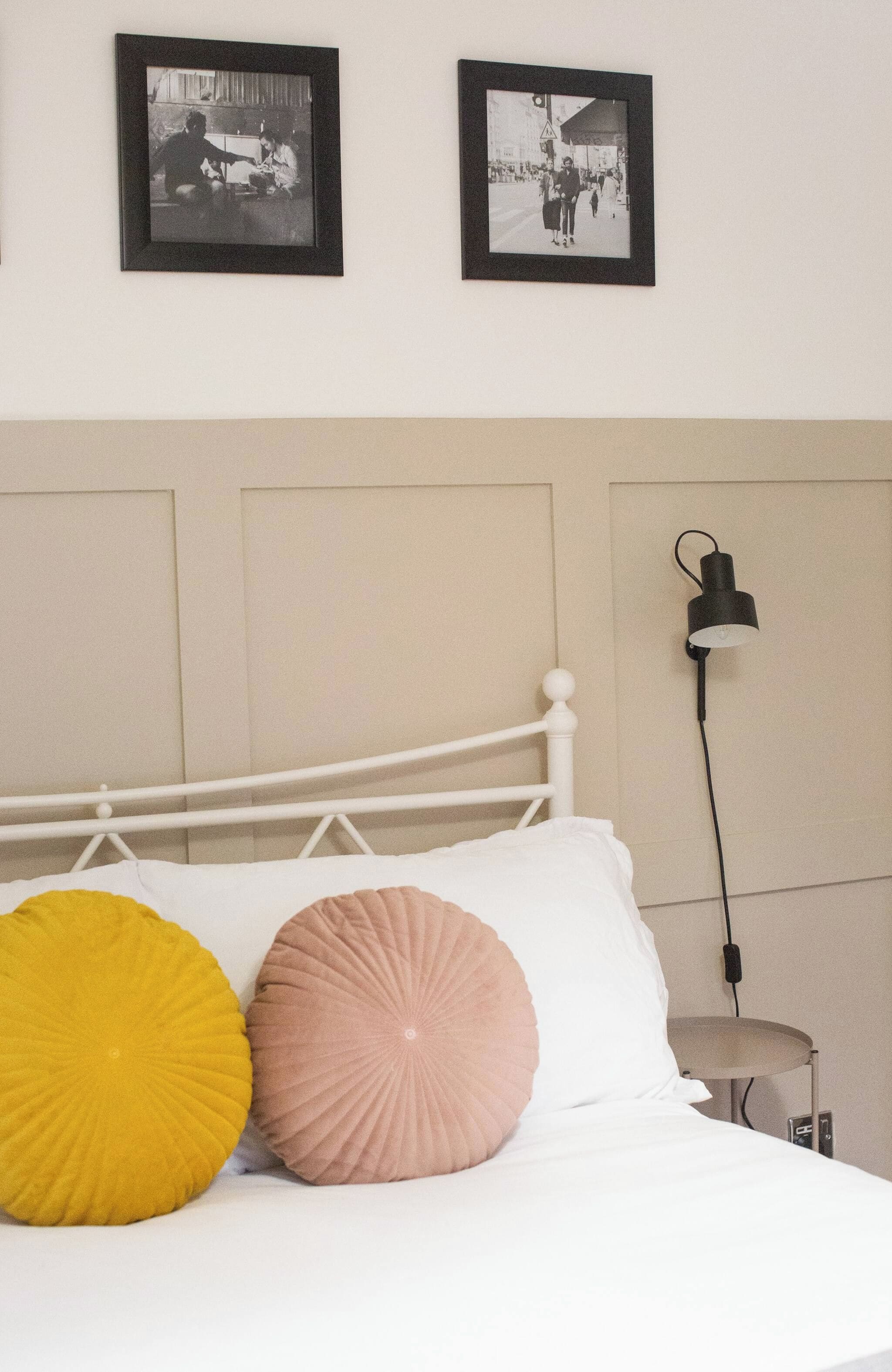 A bed with two pillows, a wall-mounted lamp, and two framed pictures above it.