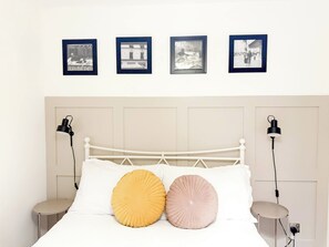 Un lit avec une tête de lit blanche, deux lampes de chevet et deux oreillers ronds, un jaune et un rose, dans une pièce où sont accrochées quatre images encadrées au mur.