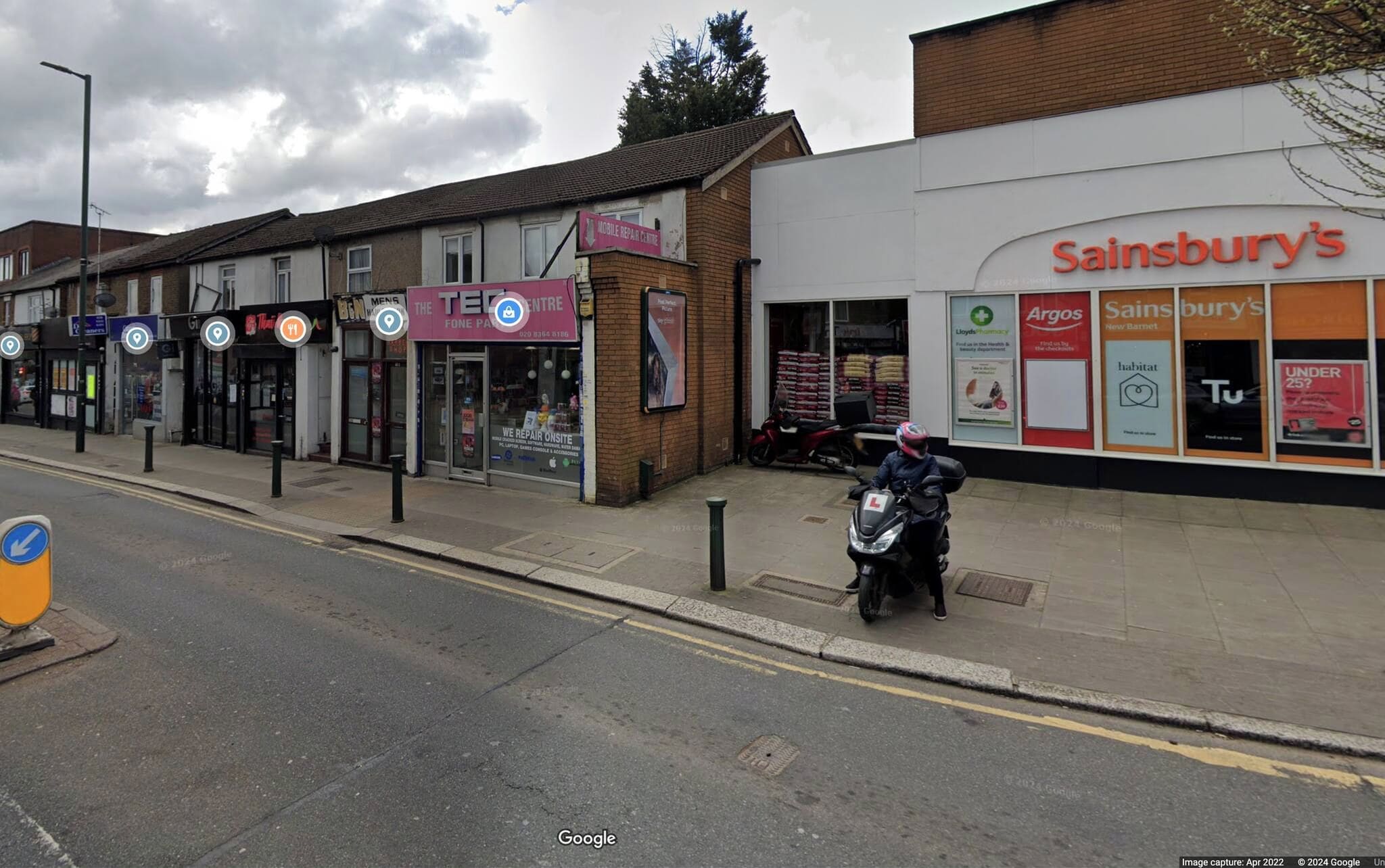 Vista de la calle con una tienda Sainsbury's, un centro de reparación y una motocicleta estacionada en la acera.
