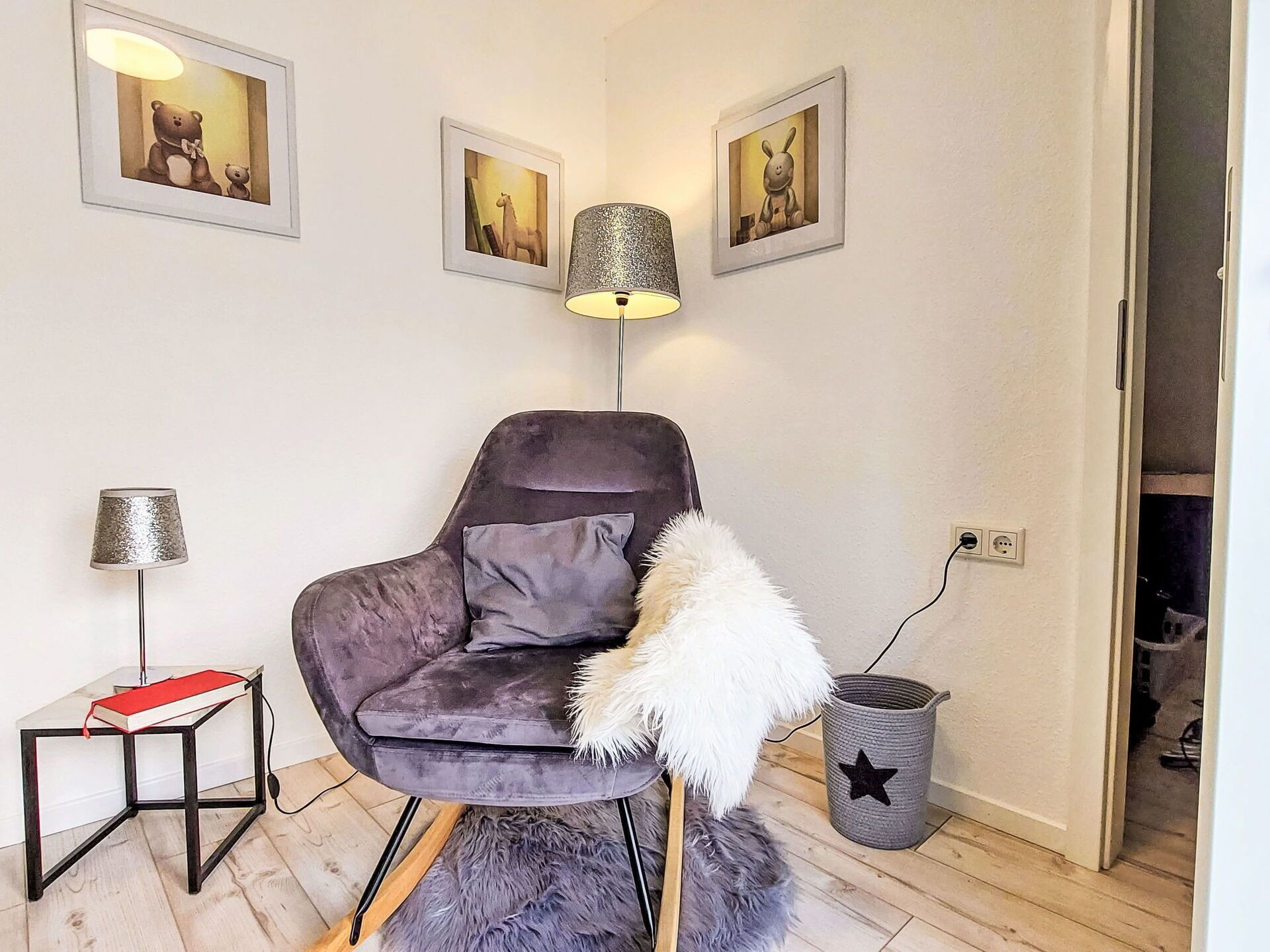 Une pièce moderne avec un fauteuil de couleur gris foncé, une petite table d’appoint sur laquelle se trouve un livre, une lampe, et des tableaux encadrés accrochés au mur.