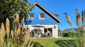 Une maison bleue avec un patio, une antenne parabolique et une pelouse où poussent de hautes herbes.