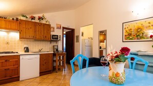 A compact kitchen with wooden cabinets, a microwave, and a dishwasher. A dining table with chairs is in the foreground, and a vase with flowers is on the table. - Beautiful apartment for 6 people with A/C, WIFI, pool, patio, pets allowed and panoramic view (Civezza)