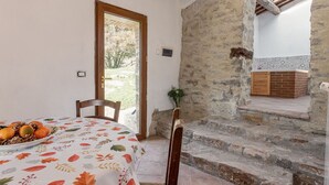 Une pièce rustique avec un mur en pierre, une table en bois sur laquelle est posé un panier de fruits, et une vue sur l’extérieur par une porte.