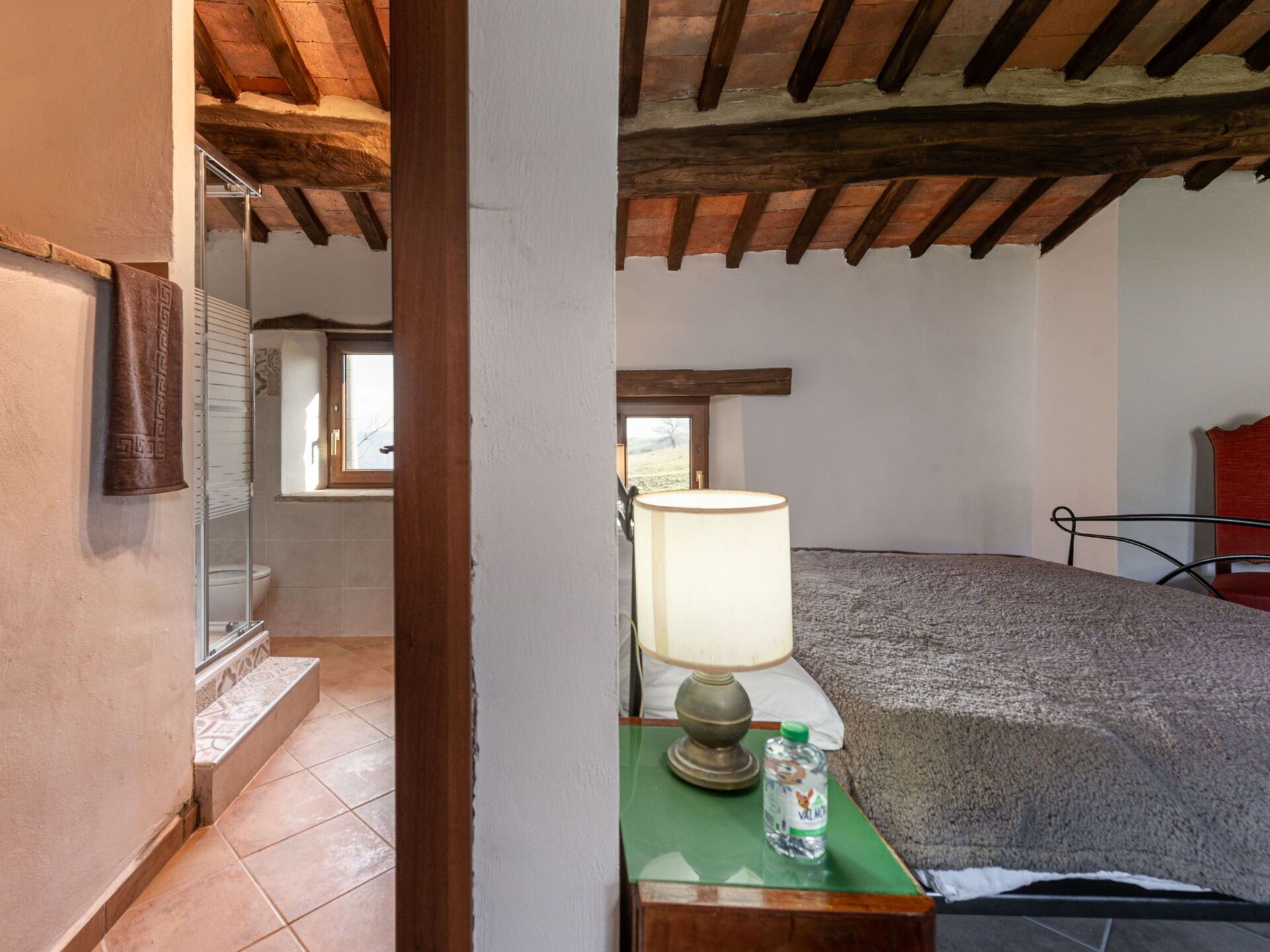 A bedroom with a wooden ceiling, a bed, a nightstand with a lamp, and a window with a view of the outdoors.