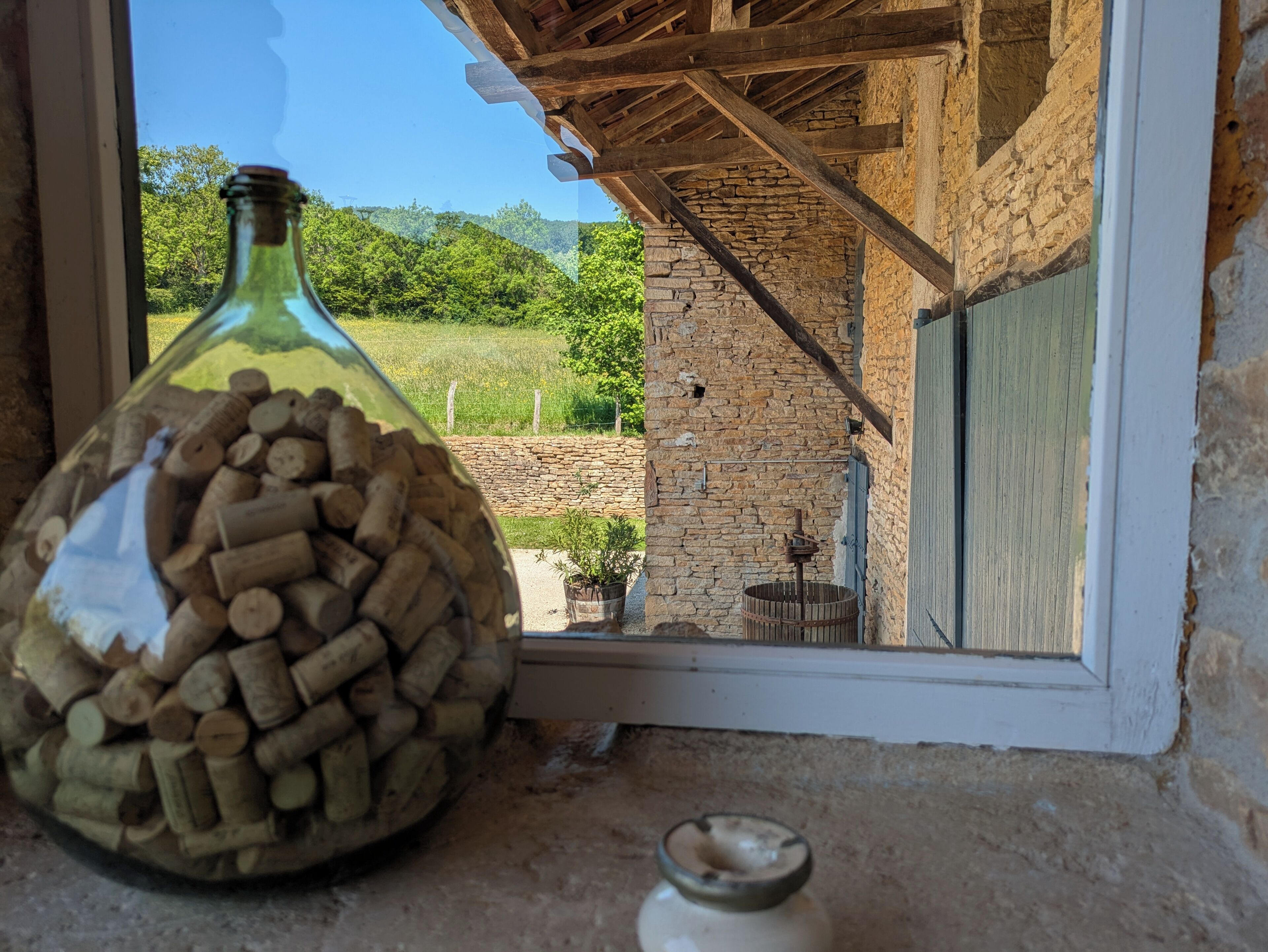 A bottle filled with corks on a windowsill, with a view of a stone building and greenery outside.