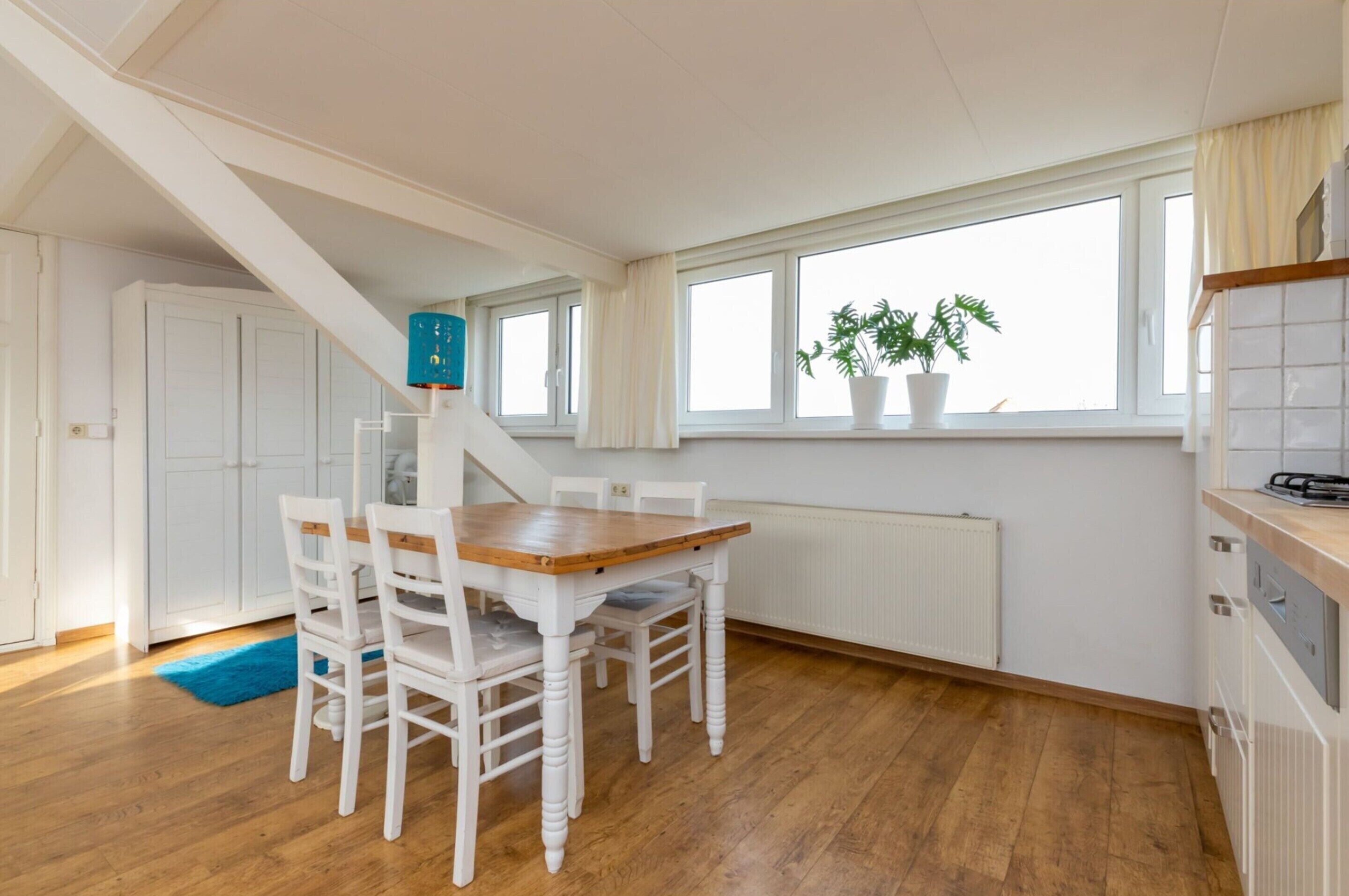A dining area with a wooden table and white chairs, a staircase, a window with a plant, and a radiator.