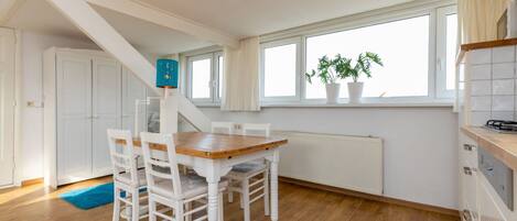 A dining area with a wooden table and white chairs, a staircase, a window with a plant, and a radiator.