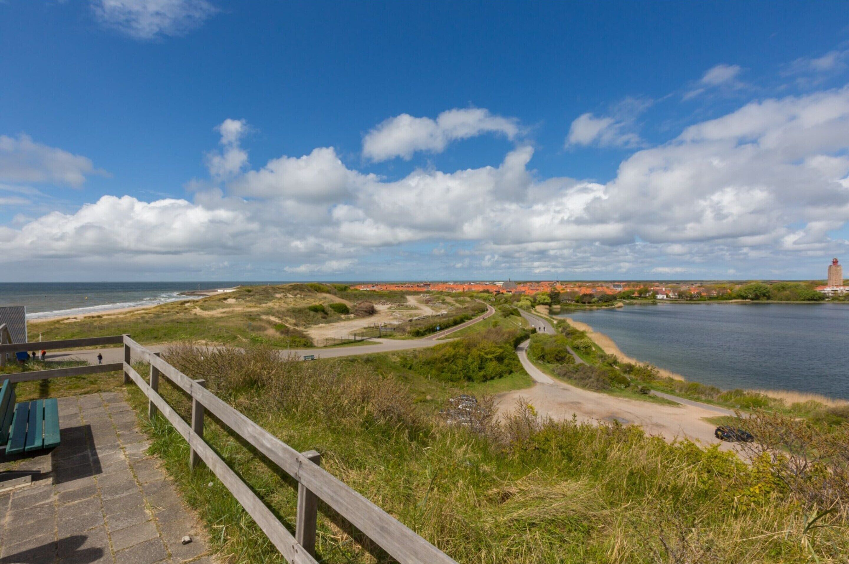 A coastal landscape with a wooden walkway, a bench, and a distant view of a town and a lighthouse.