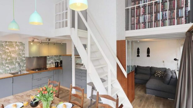 Un amplio salón con una escalera que sube a un altillo, una mesa de comedor dispuesta para el desayuno y una estantería llena de libros.