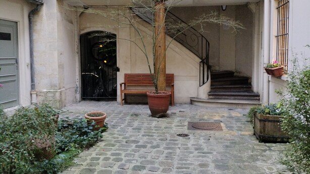 A courtyard with a cobblestone path, a wooden bench, a potted tree, and a staircase leading to an upper level.